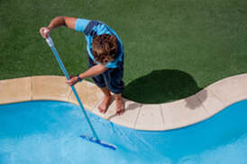 Picture of a man using a pool brush to clean the sides of a pool.  He is standing on the edge of the pool brushing down into the pool.